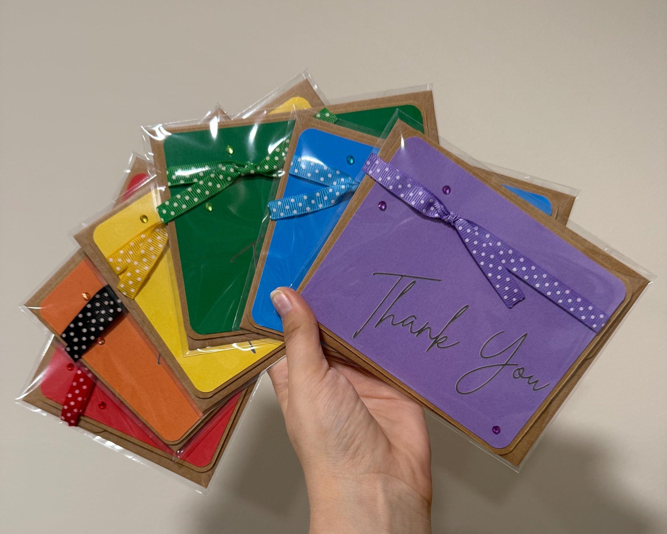 Hand holding a purple 'Thank You' card among other colorful cards on a beige background