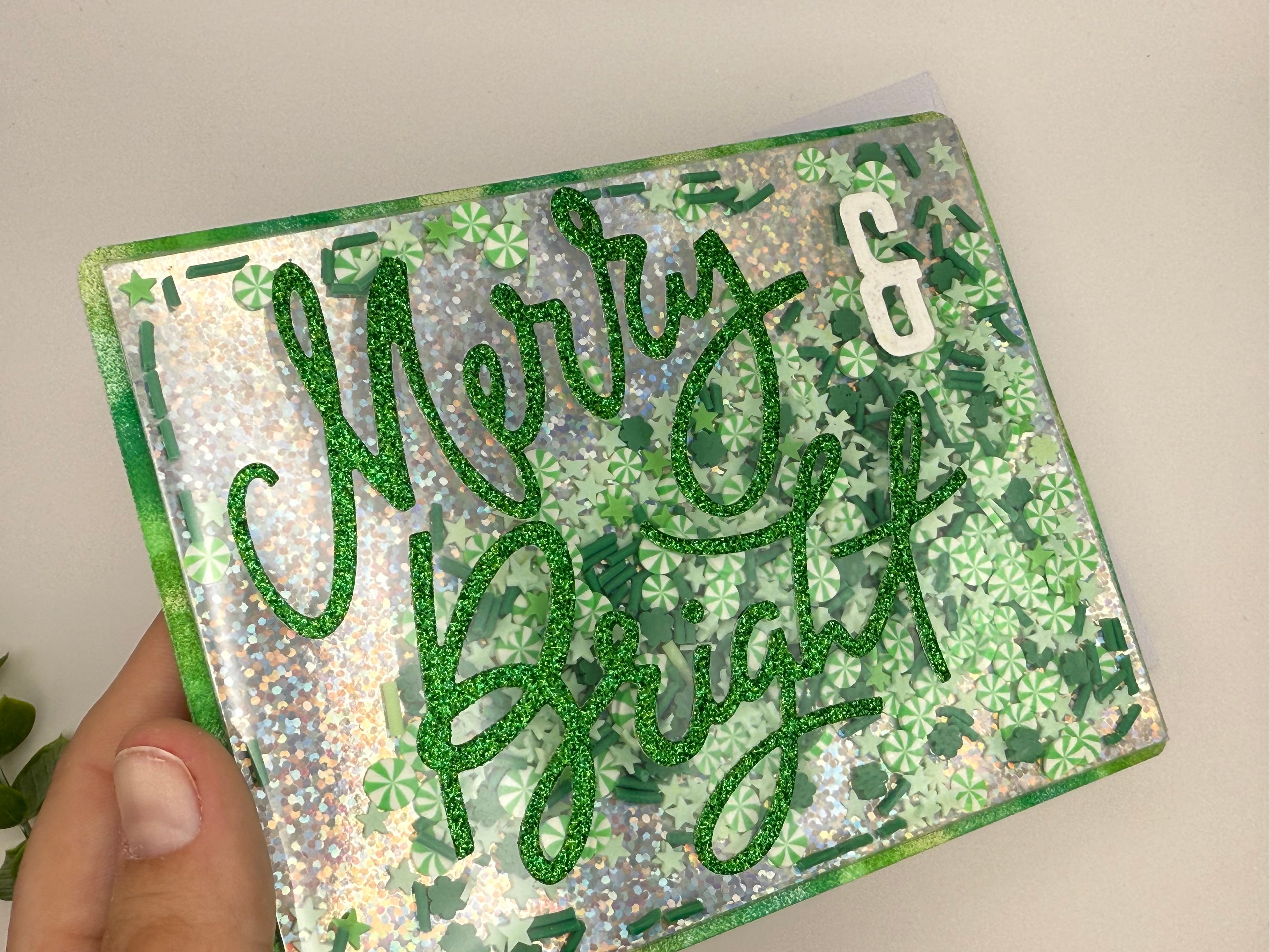 Hand holding a card with 'Merry Bright' text in green glitter on a floral patterned background.