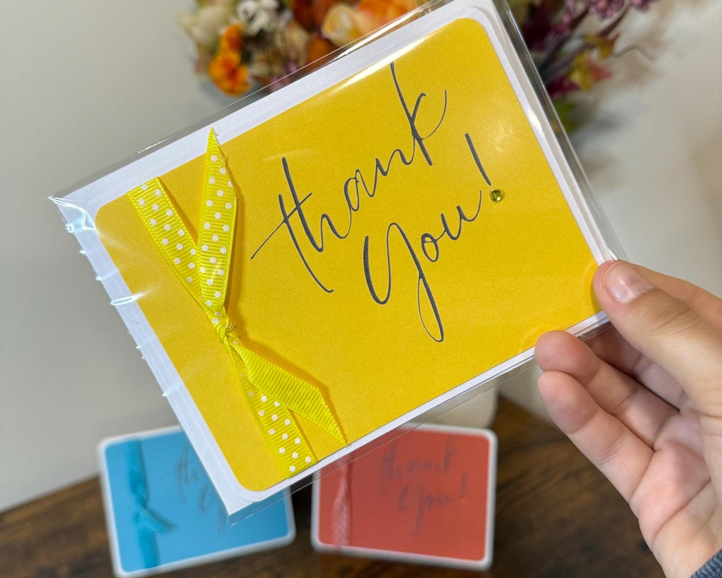 Yellow 'thank you!' card held by a hand with flowers in the background