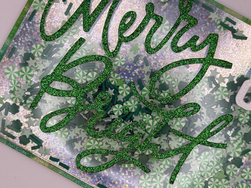 Green glittery 'Merry Christmas' text on a decorative card with floral patterns.