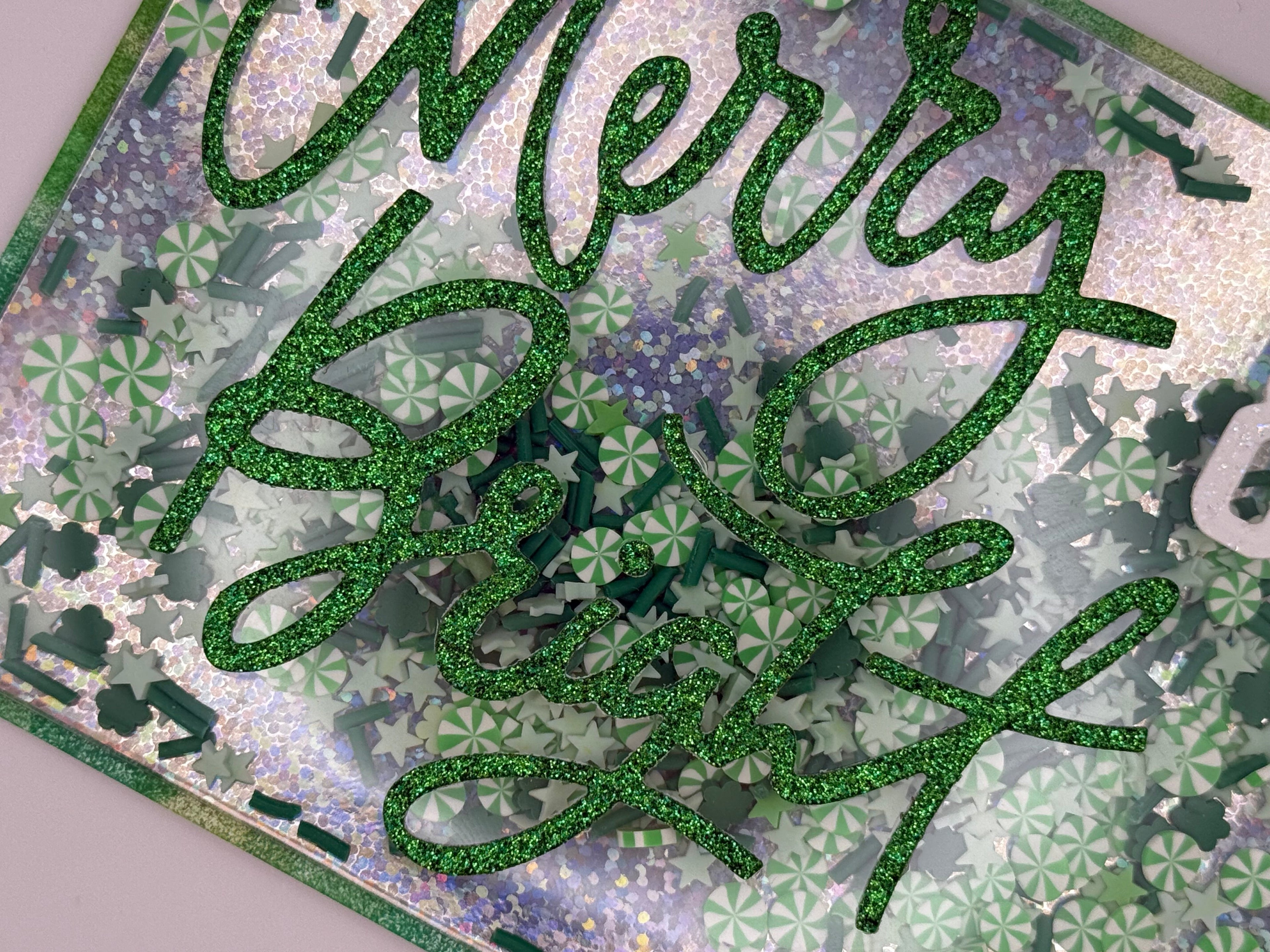 Green glittery 'Merry Christmas' text on a decorative card with floral patterns.